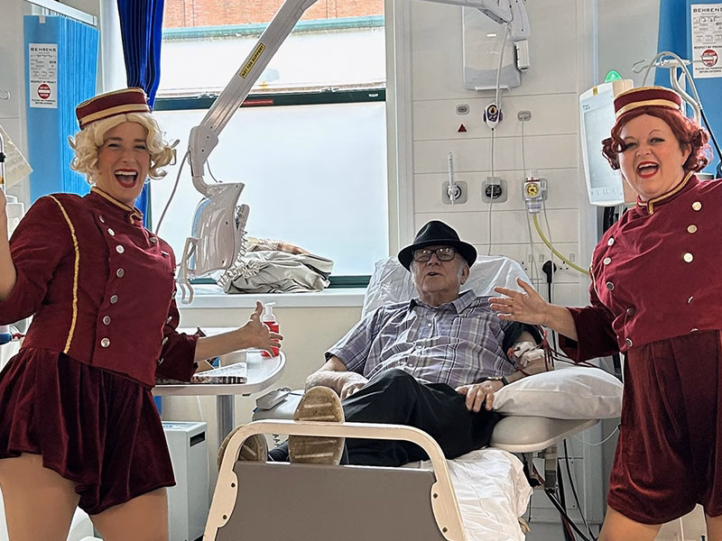 2 performers smiling at the camera, standing around a patient in a hospital bed