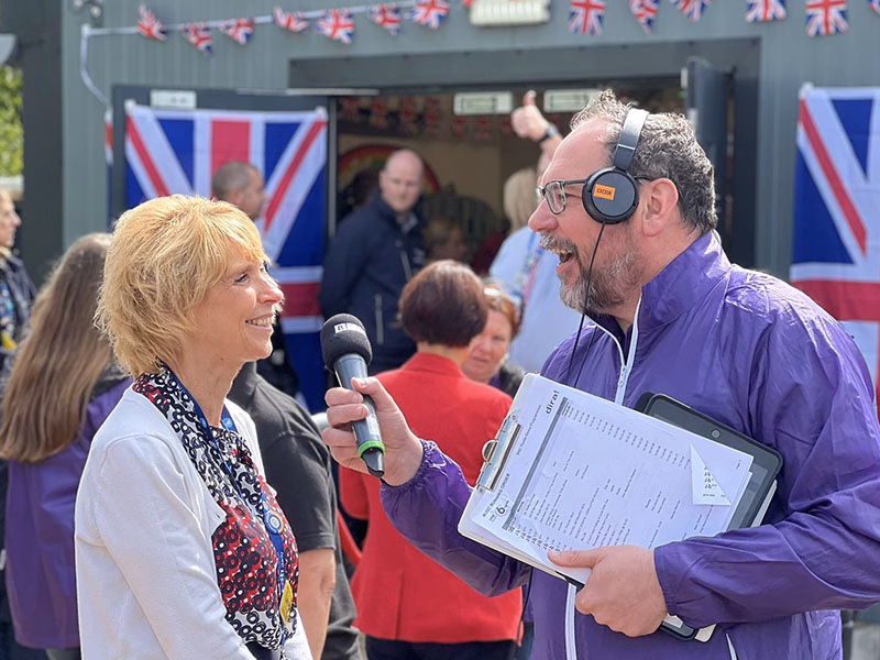 2 people smiling outdoors. One is holding a microphone towards the other interviewing for BBC Radio